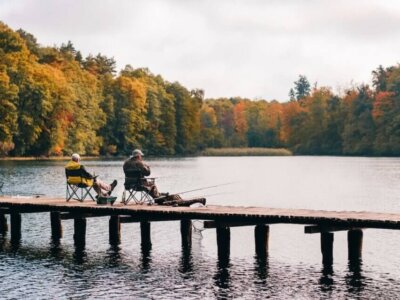 Twee mensen op een pier, aan het vissen. Op de achtergrond een meer en bos in de herfst.